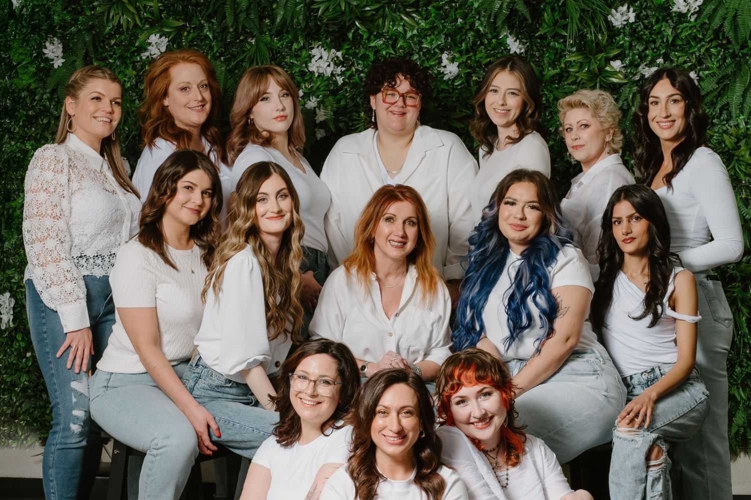 Group of women in white tops and jeans posing in front of green foliage backdrop.