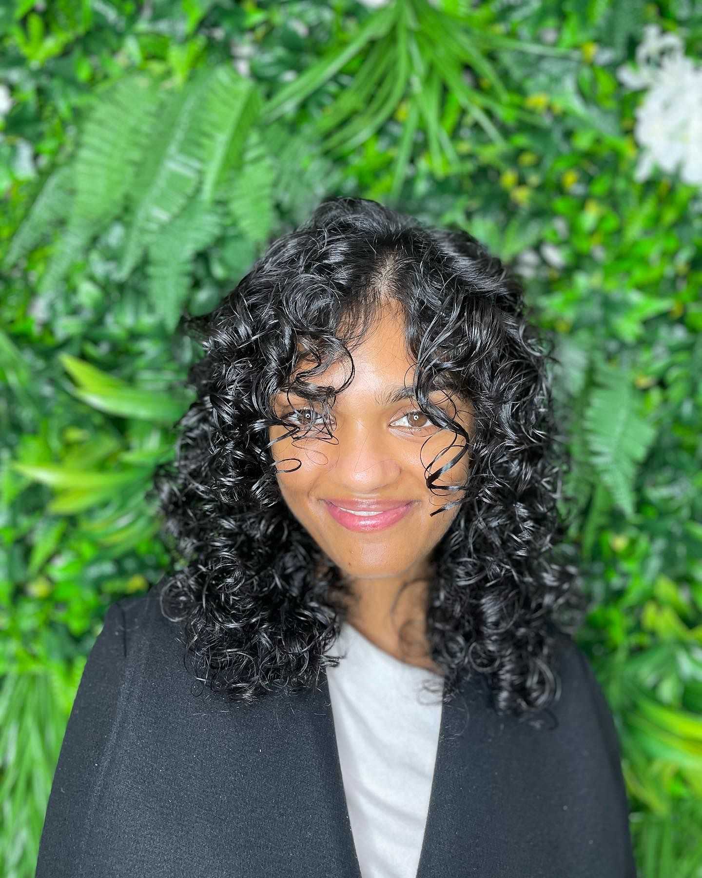 Smiling person with curly hair against a vibrant green leafy background.